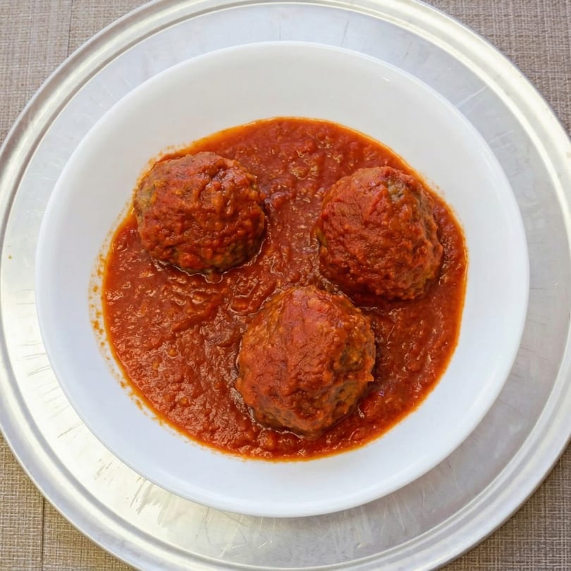 Three meatballs covered in rich red marinara sauce, served in a white bowl on a silver platter.