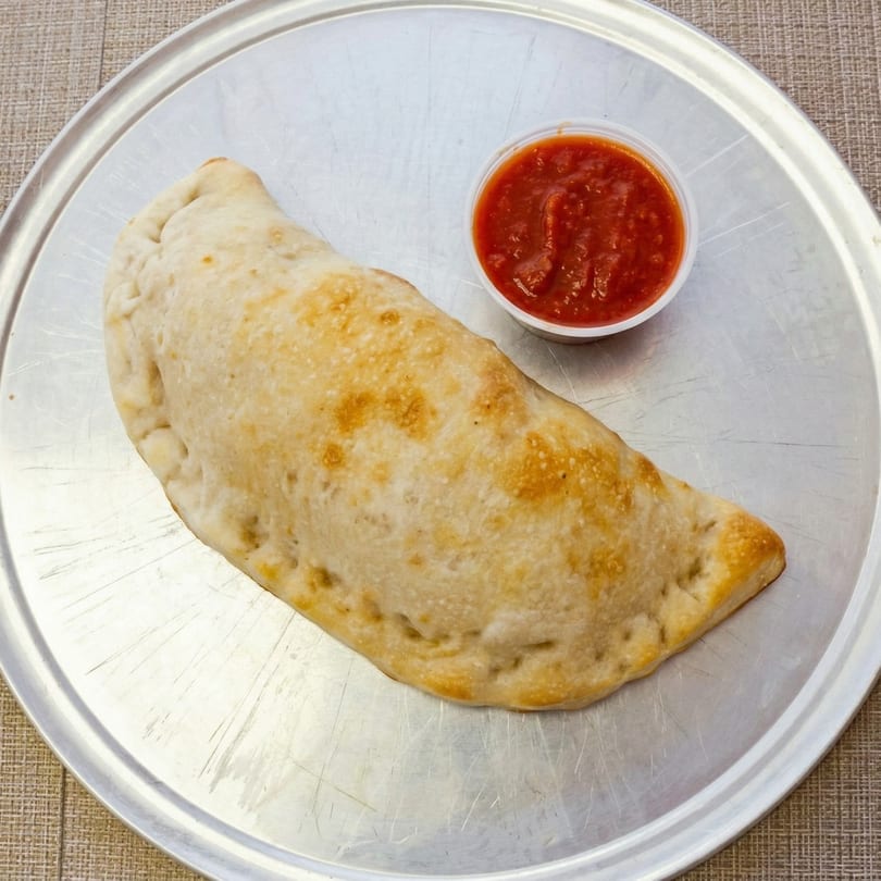 Golden-brown calzone with a side of red marinara sauce on a metal pizza pan.
