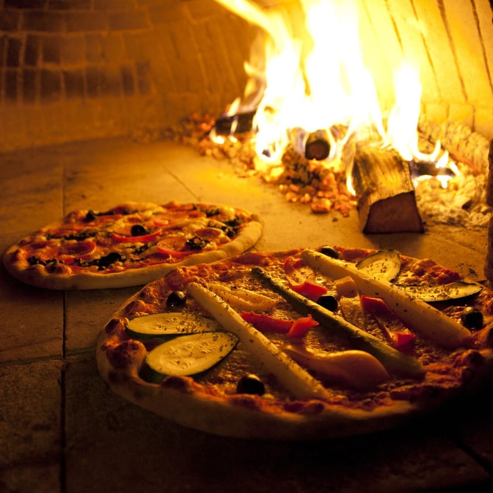 Two pizzas baking in a wood-fired oven, with flames visible in the background.